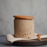 Jar of sourdough starter on wooden board with spoon and cloth wipe