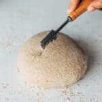 Bread dough being scored by knife held by hand