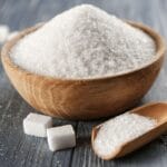 Wooden bowl of sugar on black table with spoon of sugar and scattered cubes of sugar