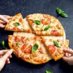 Sliced pizza on wooden pad touched by hands on black table