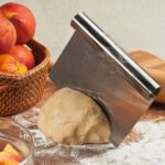 Metal dough scraper in dough with basket full of apples