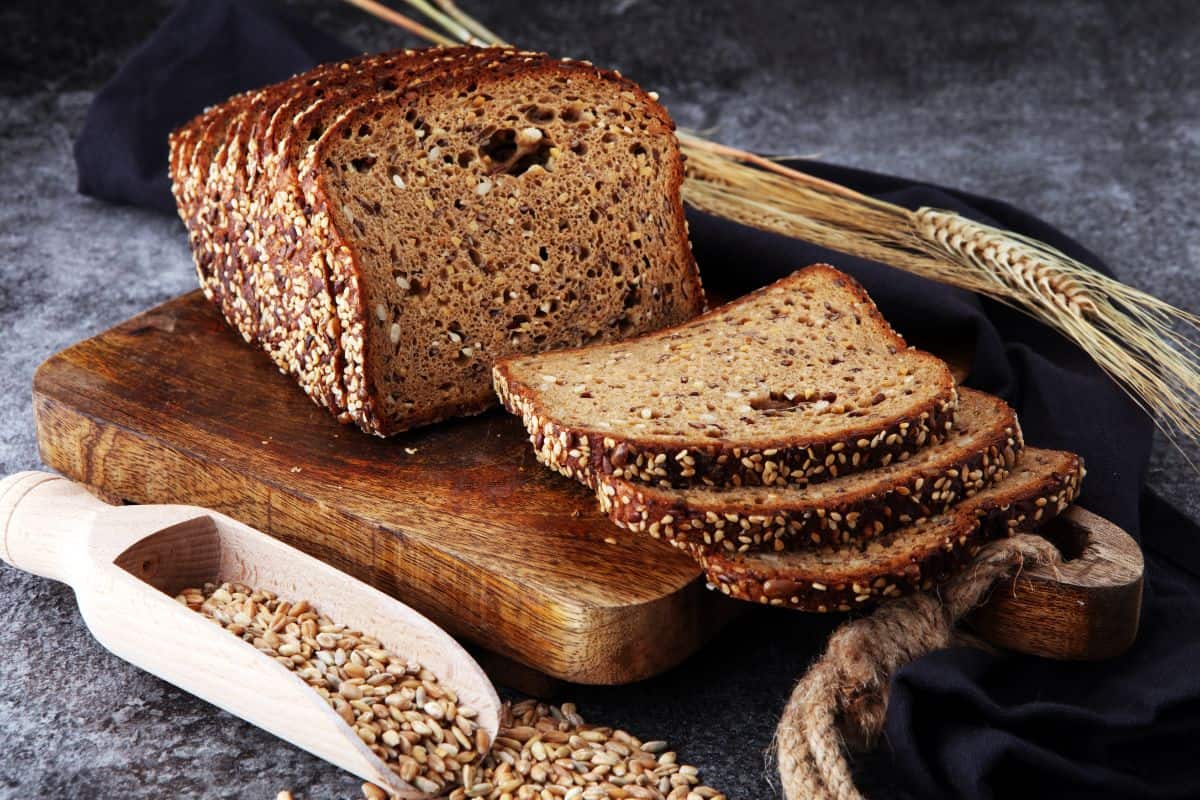 Sliced loaf of bread on wooden pad with cloth wipe, whole wheat and wheat grains on table and in wooden spatula