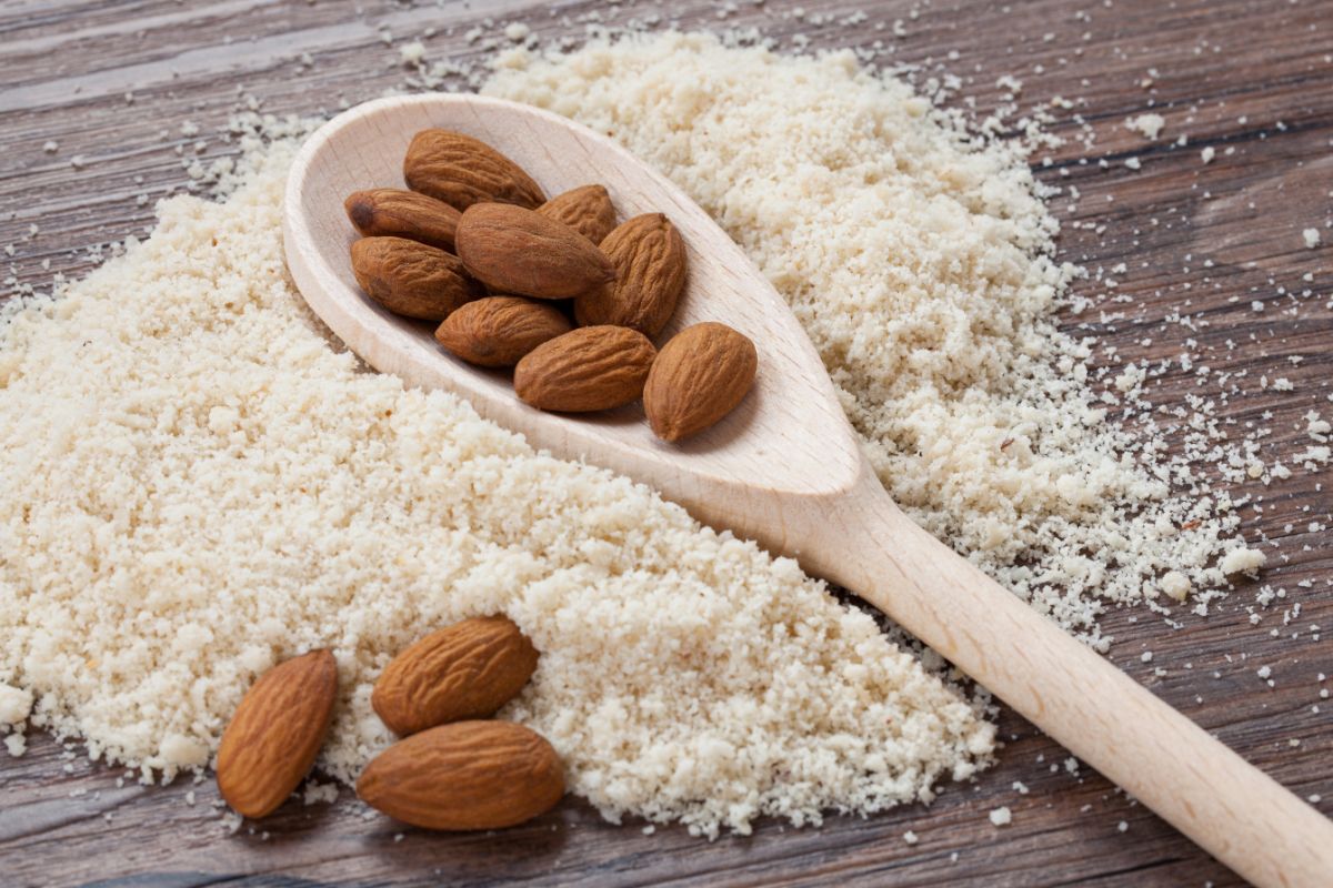 Wooden spoon full of almonds on almond flour and scattered almonds on table
