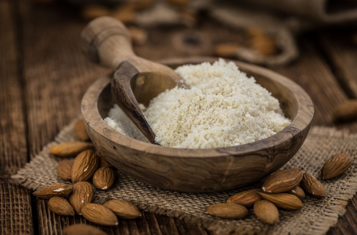 Wooden bowl of almond flour with wooden spoon on table with scattered almonds around