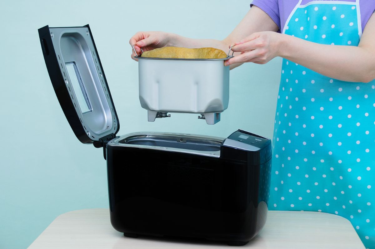 Woman holding loaf of bread in breadmaking machine pan over black breadmaker machine