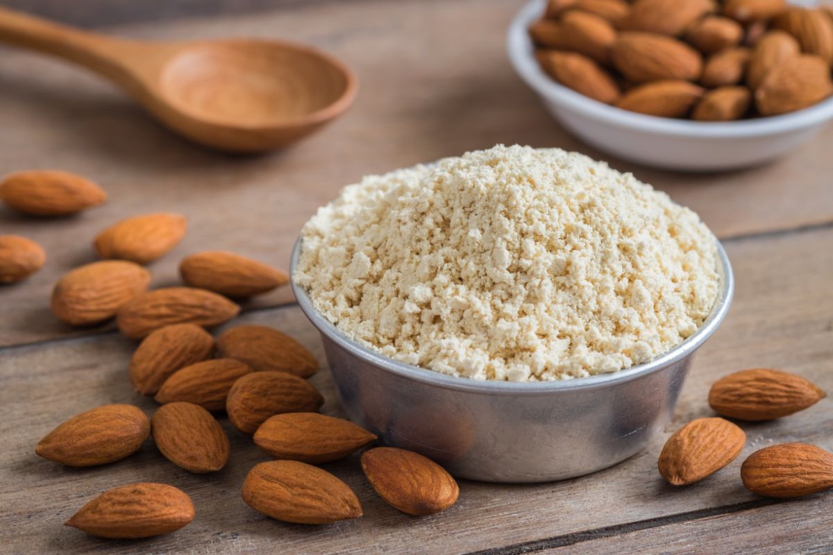 Bowl full of almond flour on table with spoon, bowl of almonds and scattered almonds around