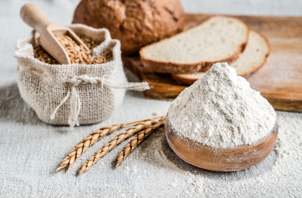 Bowl of flour next to wheat, wooden pad with bread and sack of wheat with wooden spoon on table