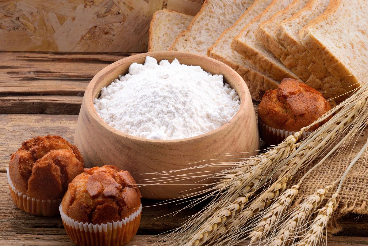 Wooden bowl of white flour on wooden table with slices of bread, muffins and stalks of wheat