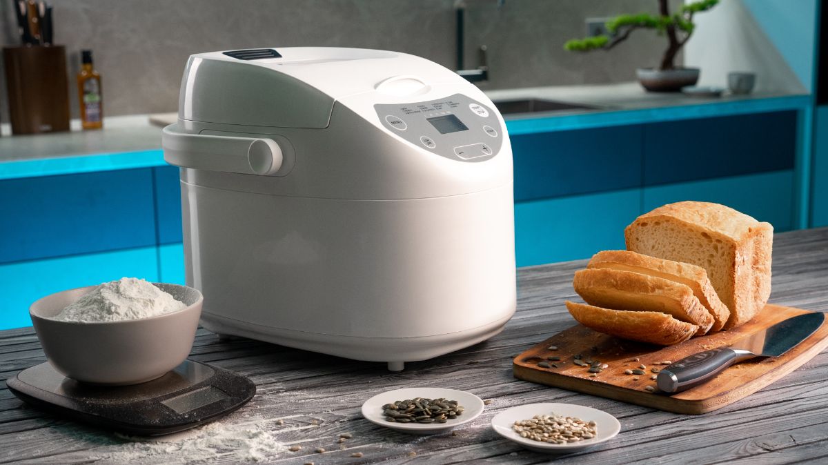 White bread maker in kitchen with small plates of ingredients, scale with bowl of flour, sliced loaf of bread on wooden pad with knife