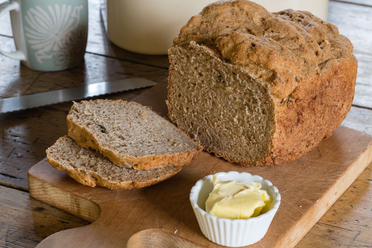 Loaf of bread partially sliced on wooden cutting board with bowl of butter