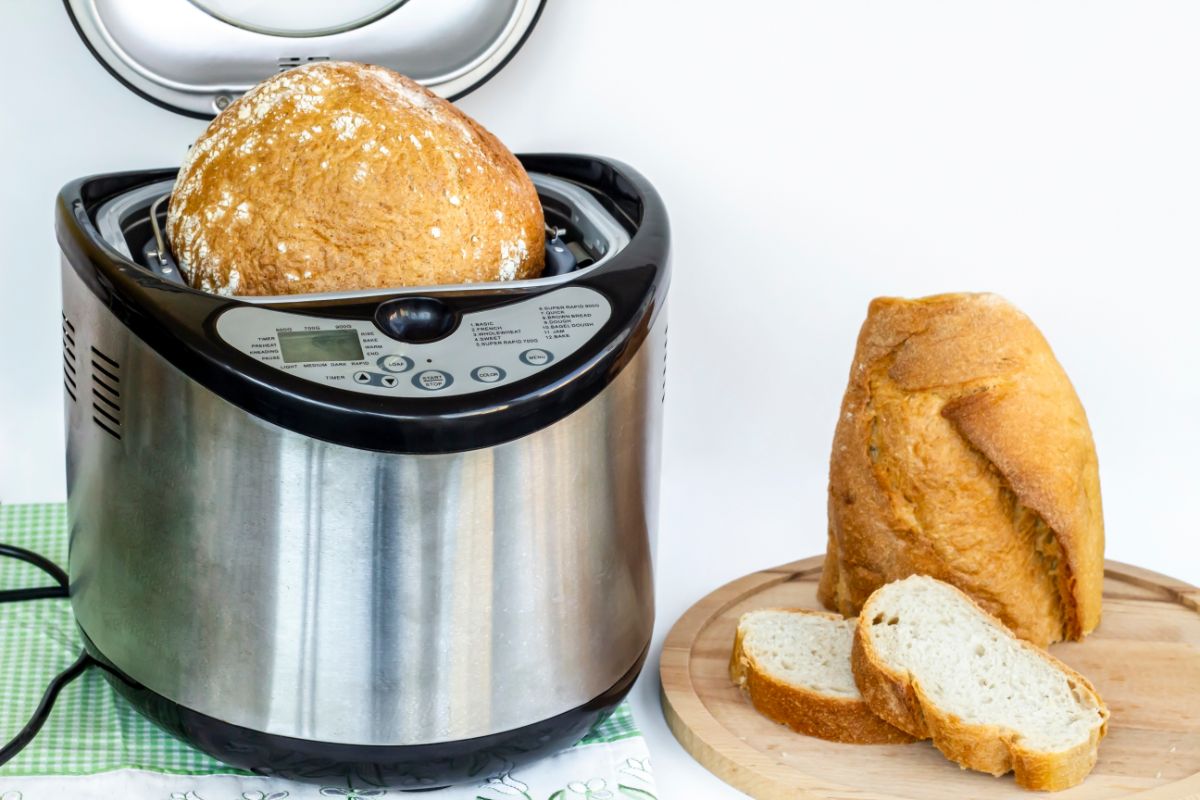 Silver breadmaker full of bread next to partialy sliced loaf of bread on wooden pad