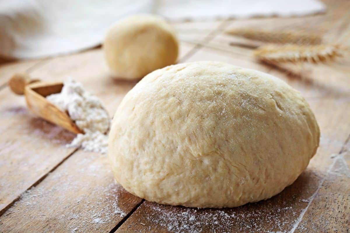 Bread dough on wooden table with wheat plant and wooden spoon full of flour in the background