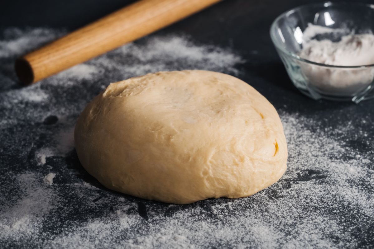 Brown dough on black table with spilled flour, flour in bowl and wooden roller