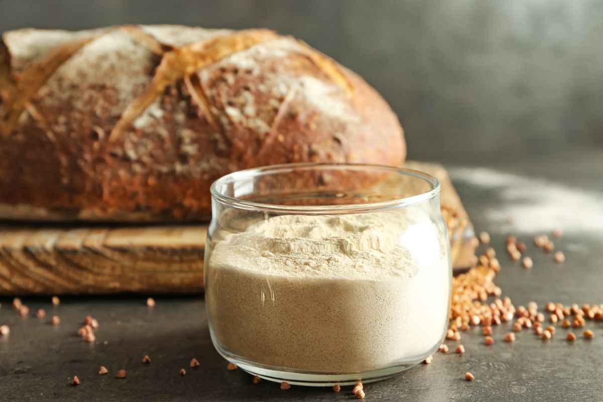 Bucketwheat flour in glass jar with loaf of  bread on cutting wooden board in the background