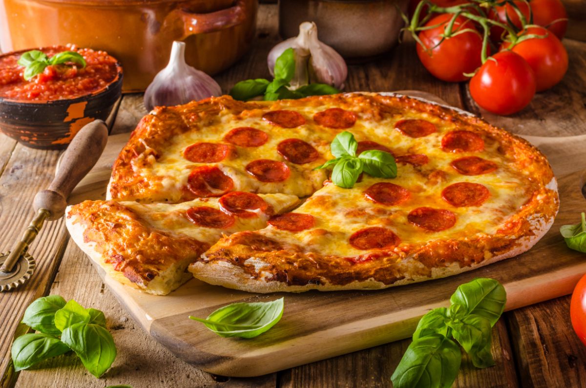Cheese pizza on wooden pad next to pizza cutter, tomatoes, herbs, garlic and bowl of sauce on wooden table