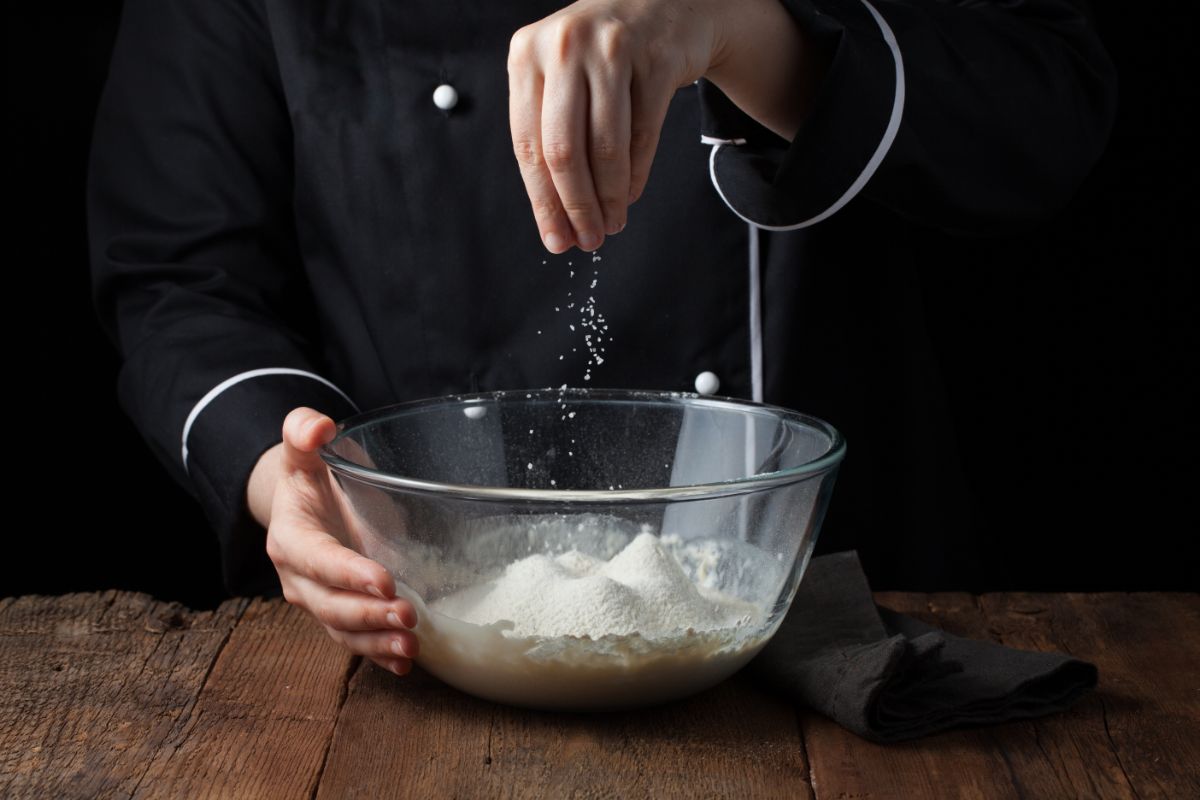 Chef sprinkling salt into glass bowl with dough on wooden table