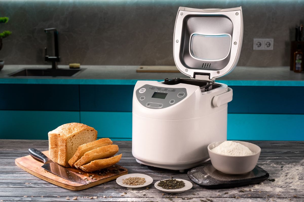 White bread maker machine on table next to scale with bowl of flour, cutting board with knife and loaf of bread and ingredients on small plates