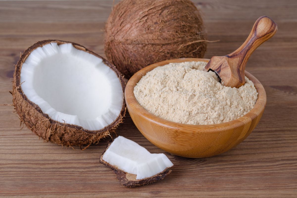Wooden bowl full of coconut flour with wooden spoon on table with coconuts