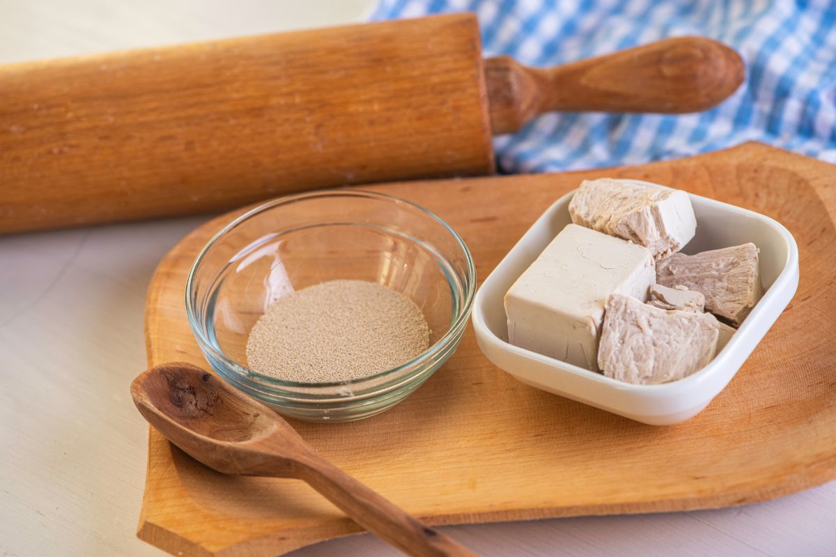 Dry and block yeast in small bowls on cuttin wooden board with wooden spoon , wooden kitchen roller and cloth wipe in the background