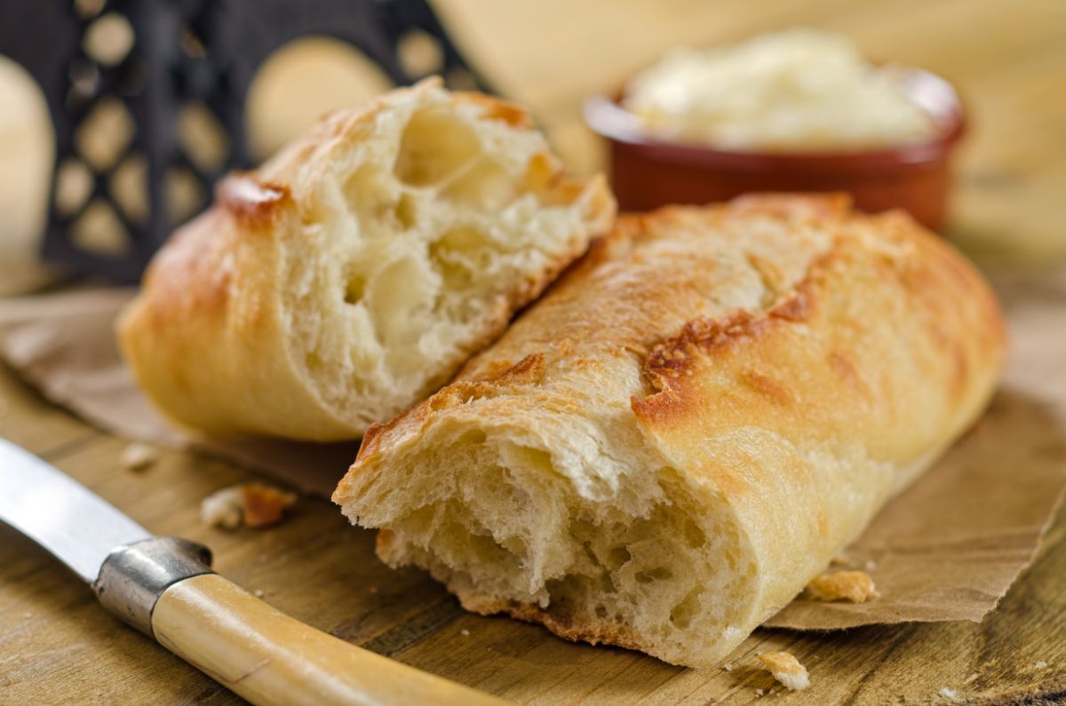French baguette on wooden pad with knife, bowl of butter in the background