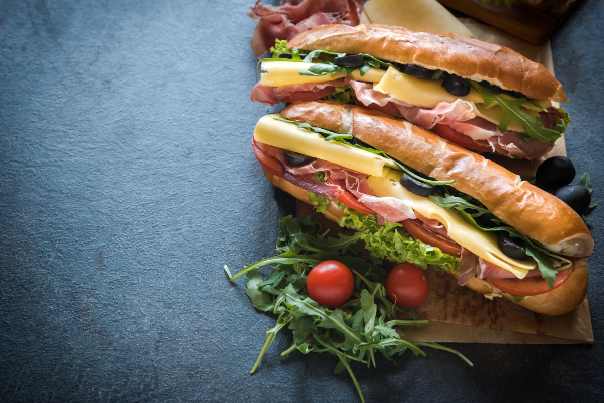 French baguettes on wooden pad with cheese, salad, tomates, bacon on black table