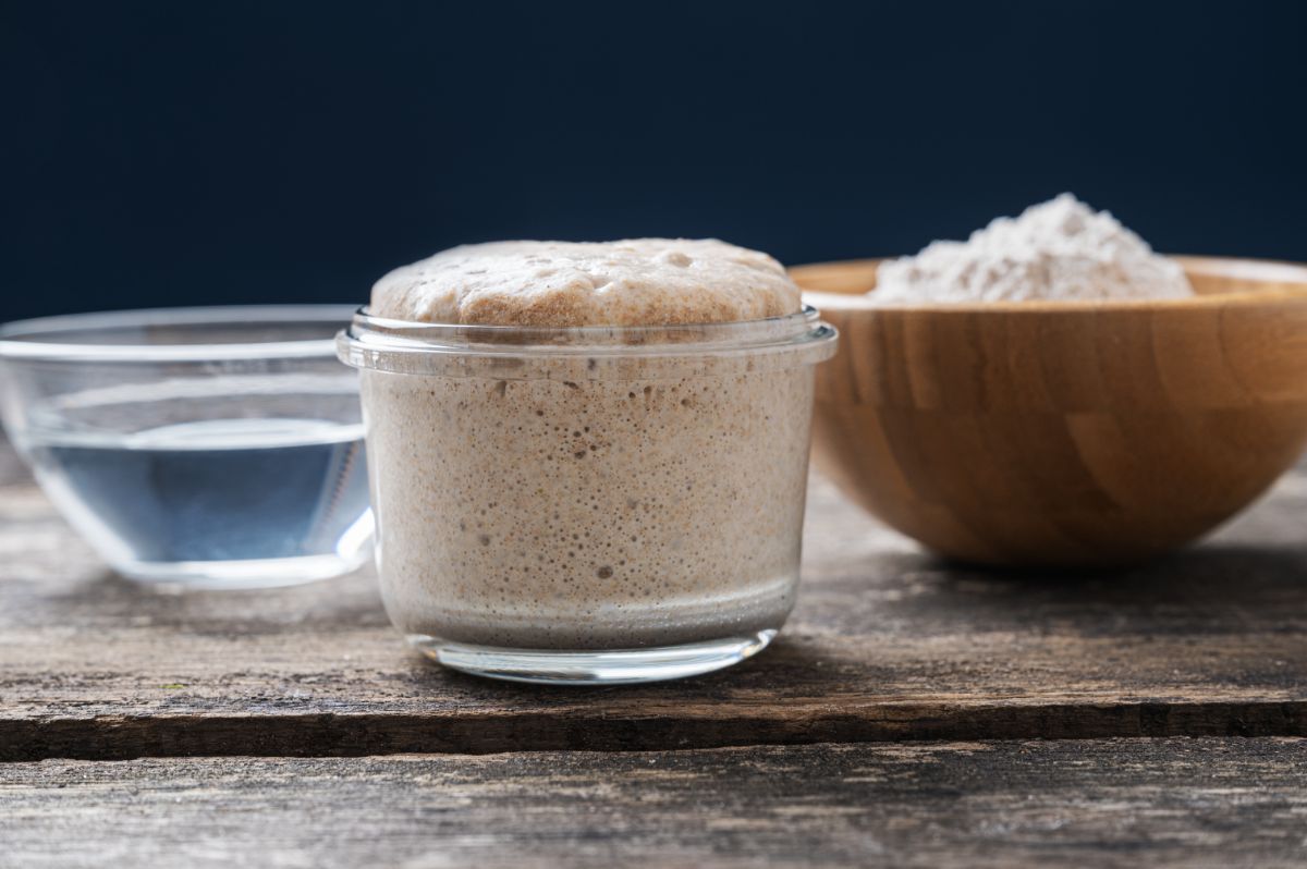 Glass jar of sourdough starter, bowl of water and bowl of dry yeast on wooden table