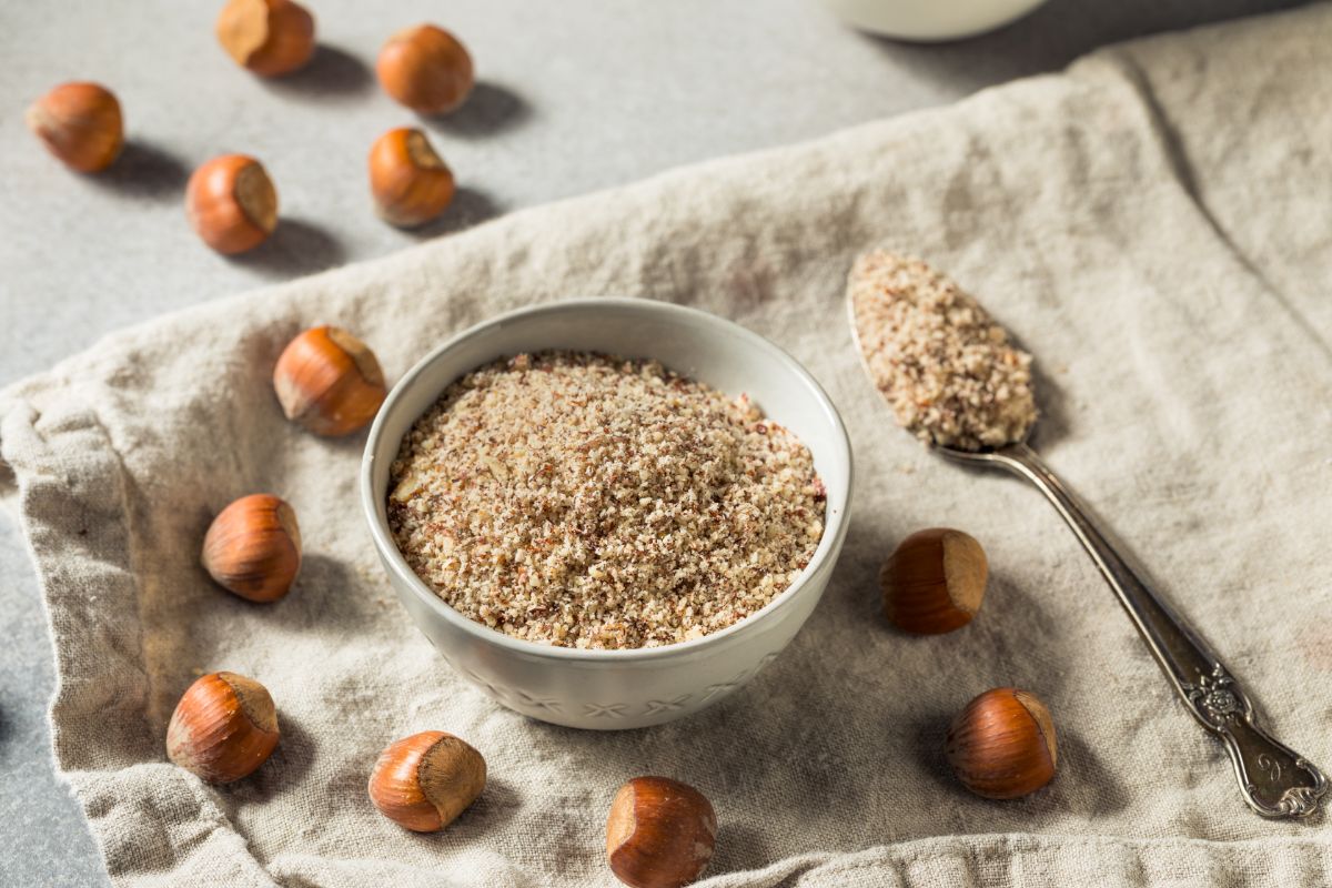 Bowl of hazelnut flour on table with spoon of flour and scattered hazelnuts around