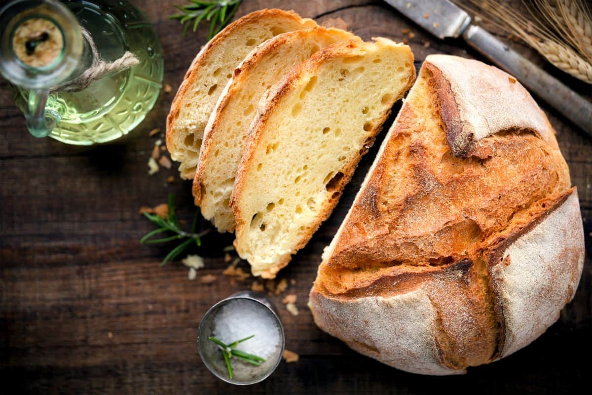 Partialy sliced loaf of bread with herbs, knife and bottle on the table