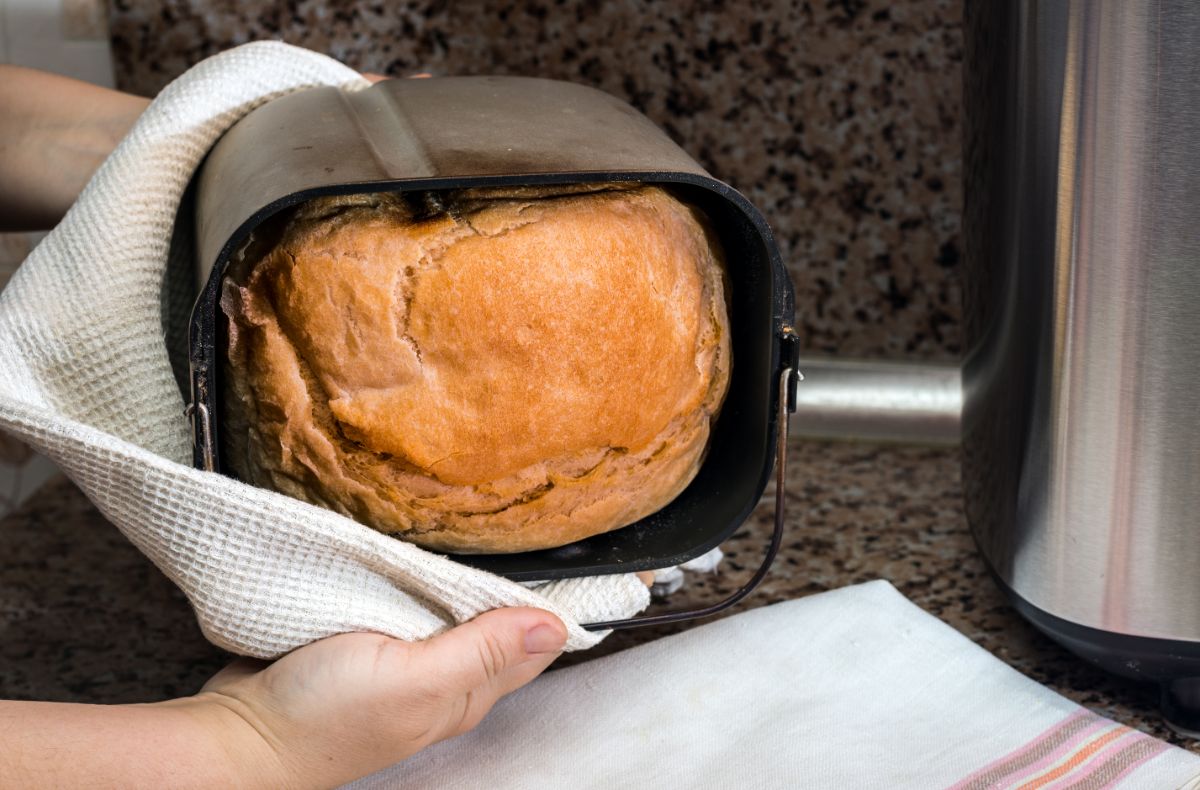 Hands holding with cloth wipe bread maker pan with baked loaf of bread over cloth wipe on table