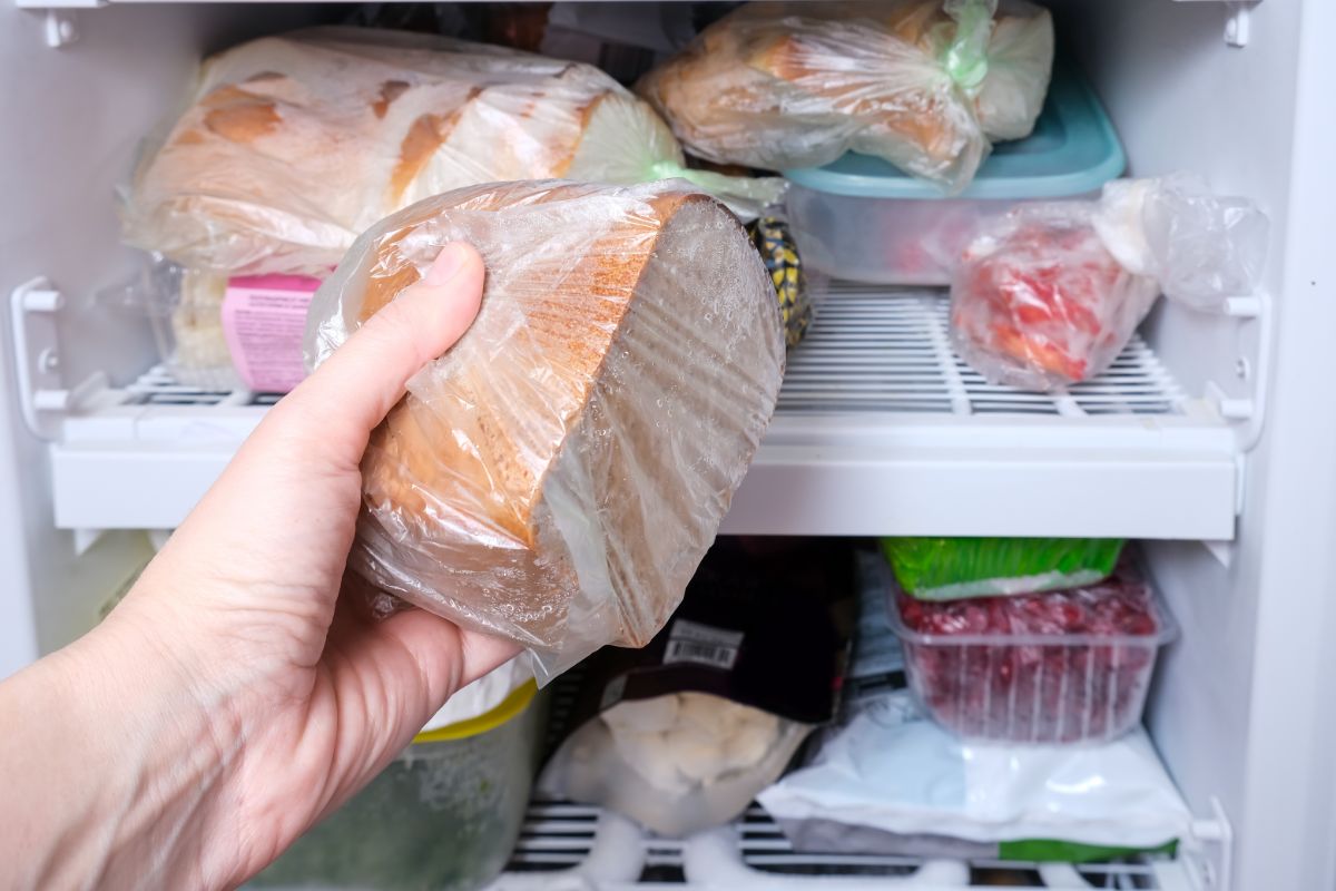 Hand holding loaf of bread in plastic bag in front of freezer full of goodies