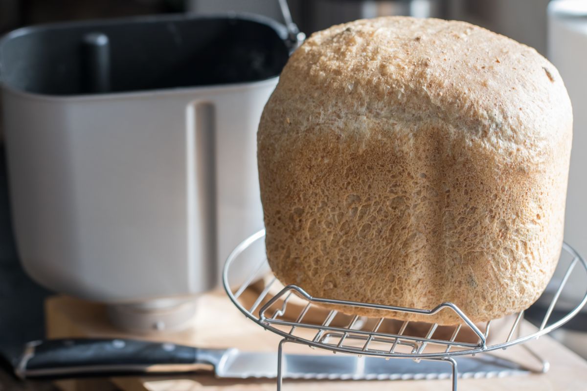 Loaf of bread on cooling rack, breadmaker pan and knife in the background