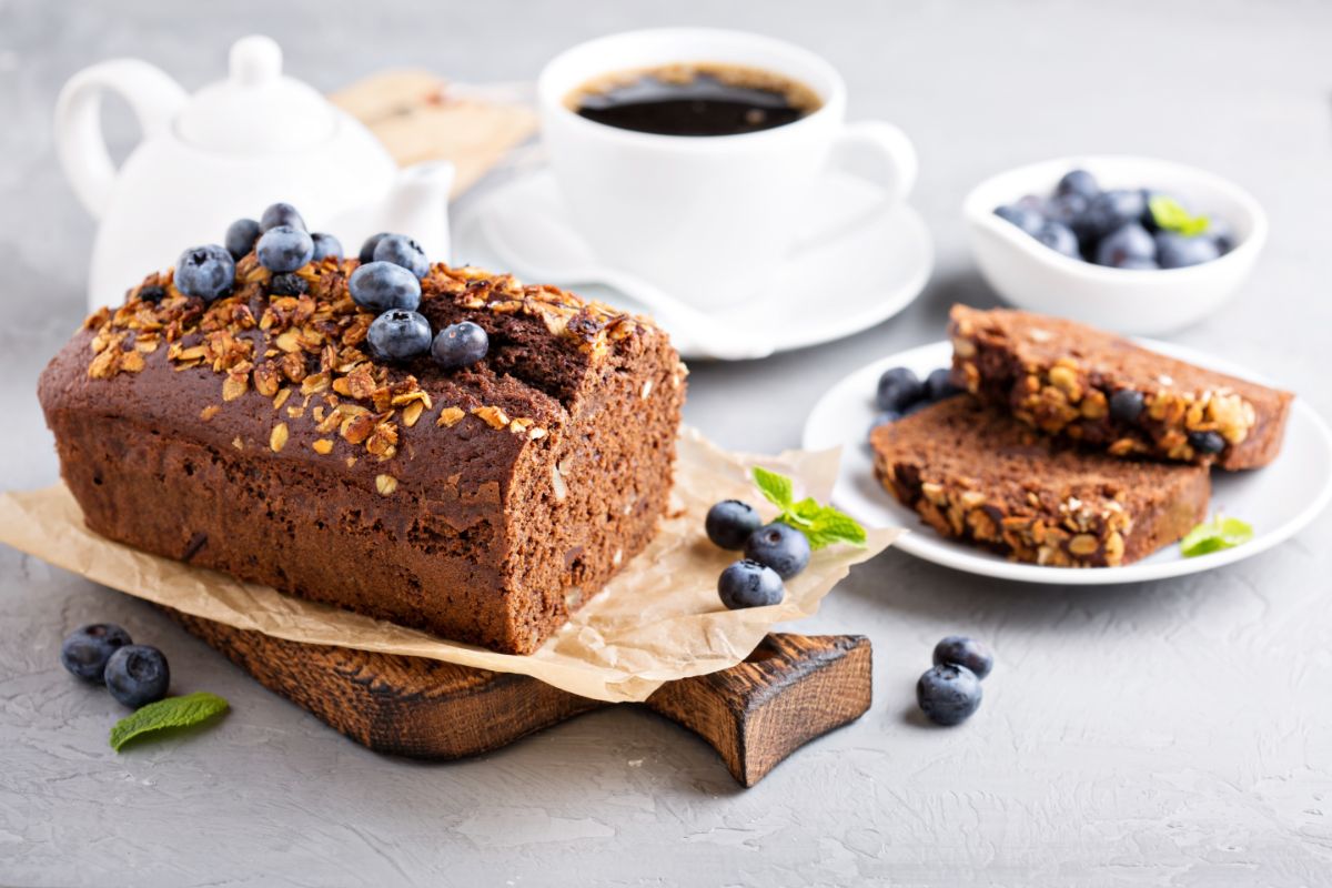 Loaf of bread on wooden pad with blueberries, slices of bread on white plate, cup of coffe, white pitcher and bowl of blueberries on the table