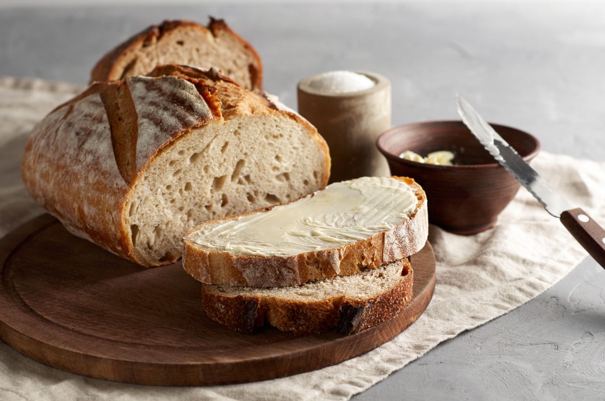 Loaf of bread with slices with butter on wooden pad next to bowl of buter with knife and salt in wooden cup