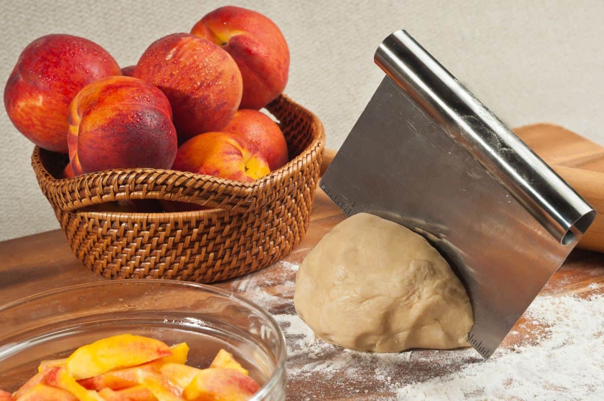 Metal dough scraper in dough with basket of apples and bowl of sliced fruits on the table
