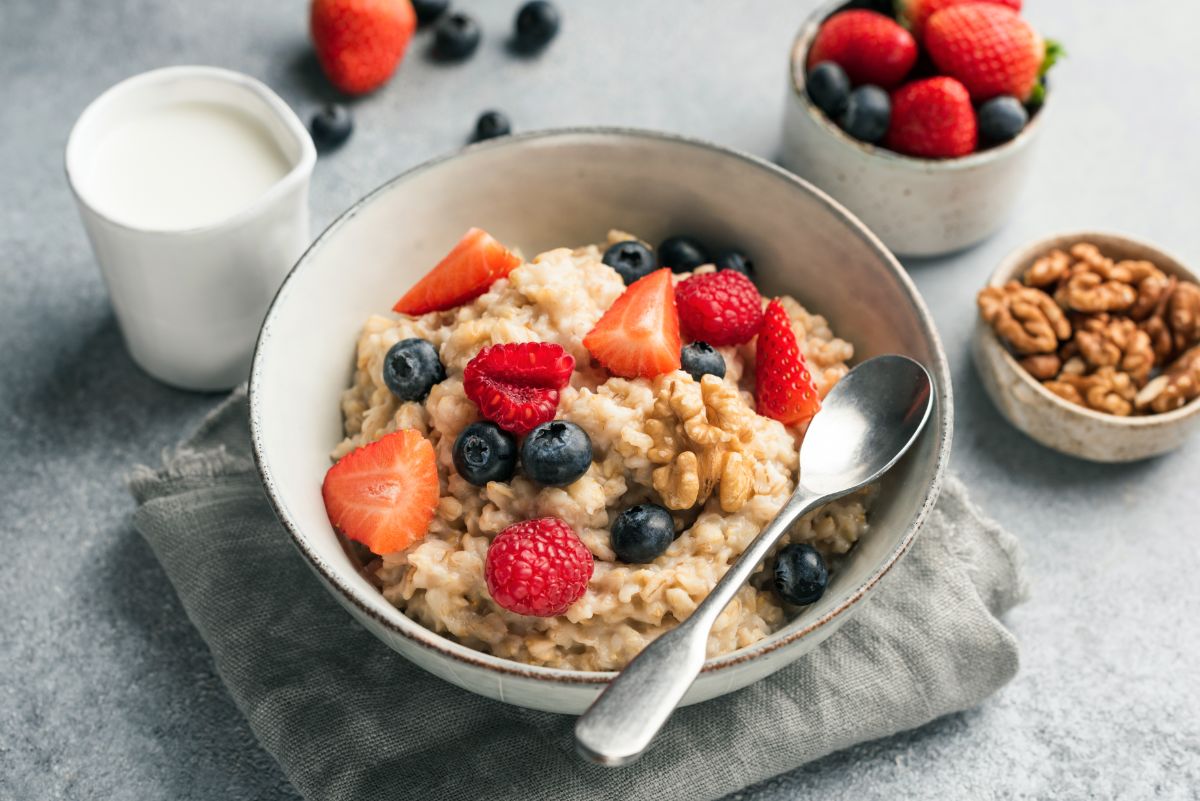 Oatmeal breakfast in white bowl with fruits and spoon on table with glass of milk, fruits in bowl and  nuts in small bowl