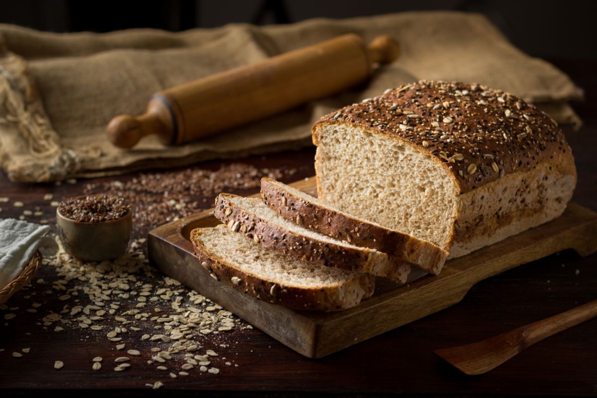 Loaf of bread partialy sliced on wooden pad with wooden roller, oat flakes, small bowl and cloth bag on the table