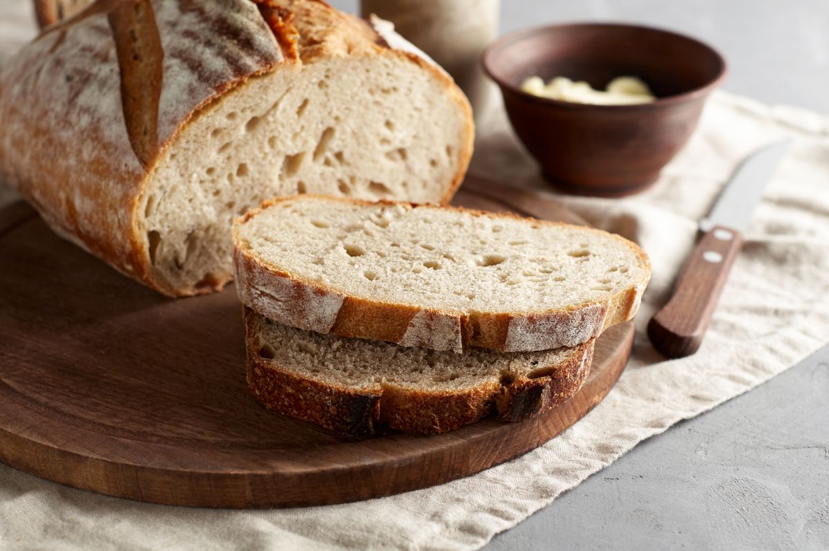 Partialy sliced sourdough loaf of bread on wooden pad next to knife and bowl of  butter