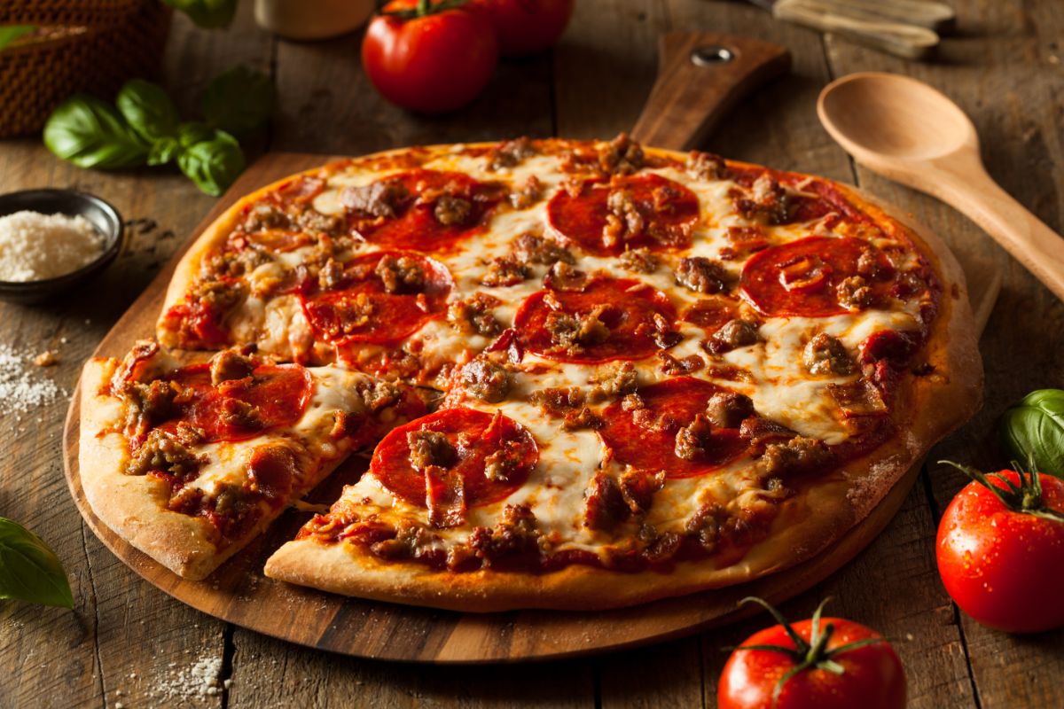 Pizza partialy sliced on wooden pad with wooden spoon, tomatoes, herbs and salt on wooden table