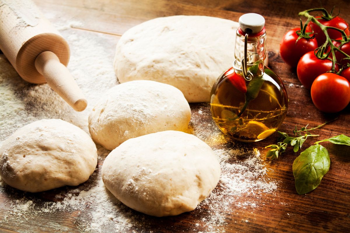 Pizza dough on wooden table with spilled flour, wooden roller, glass jar of oil and tomatoes