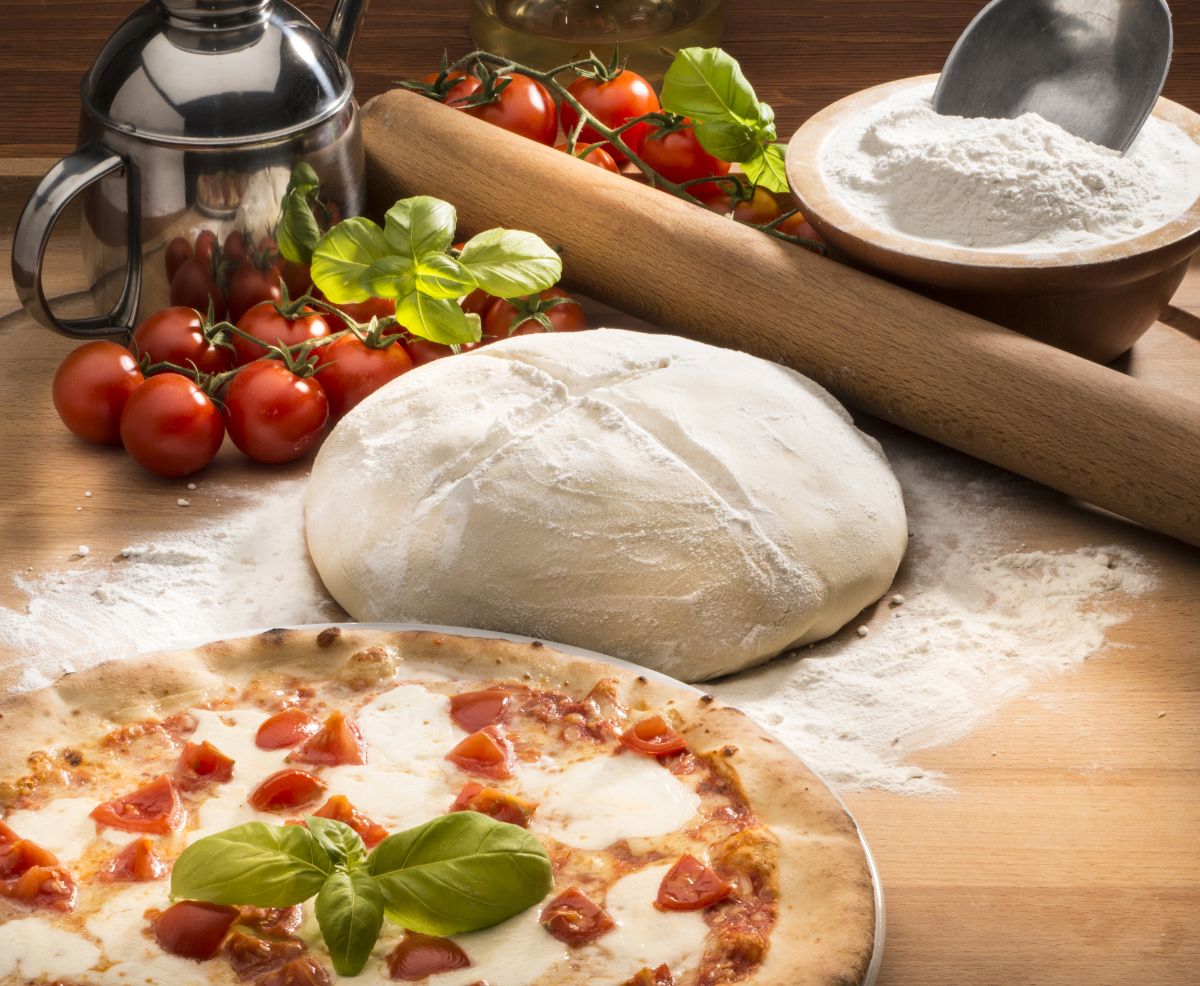 Dough, baked pizza, tomatoes, wooden roller, bowl of flour, and metal jar on wooden table