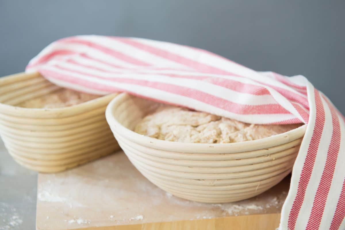 Proofing baskets with rising dough partially covered by cloth wipe on wooden board