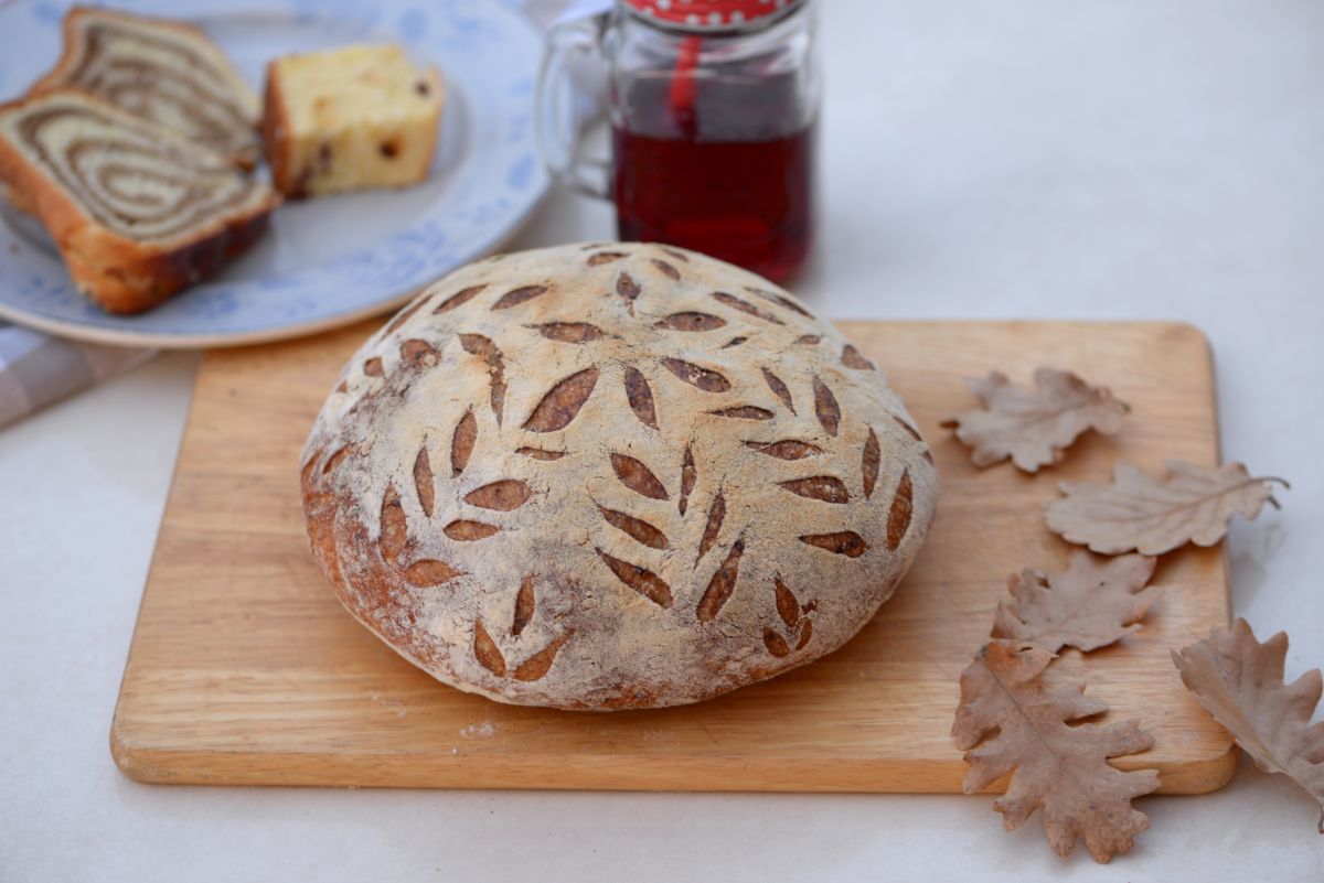 Scored loaf of bread on wooden pad on table with leaves, jar of honey and plate with pieces of pie