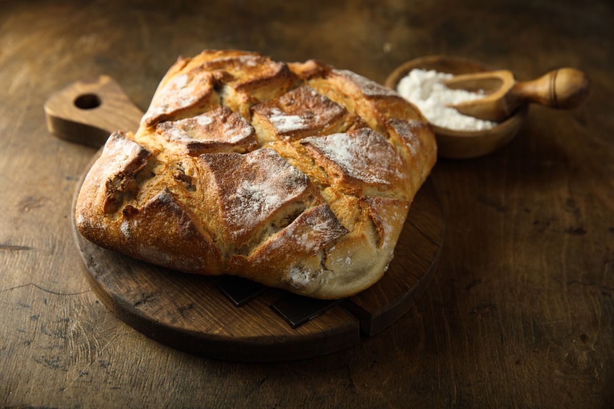 Scored sourdough loaf of bread on wooden pad next to bowl of flour with wooden spoon on wooden table