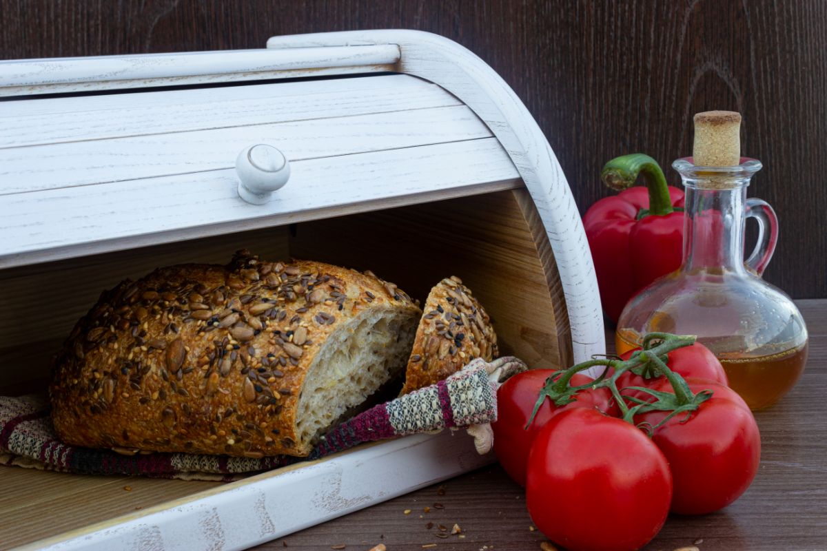 Sesame loaf of bread in wooden bread box with tomatoes and jar of oil on table