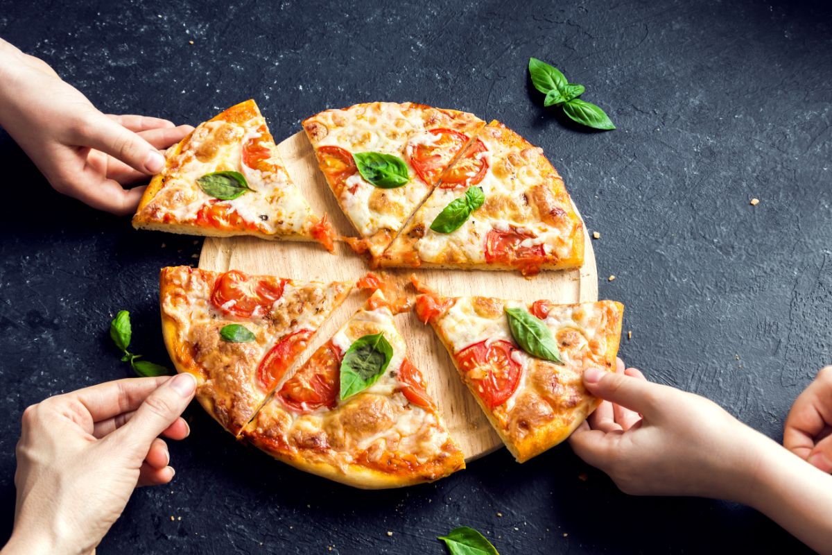 Sliced pizza on wooden pad touched by hands on black table