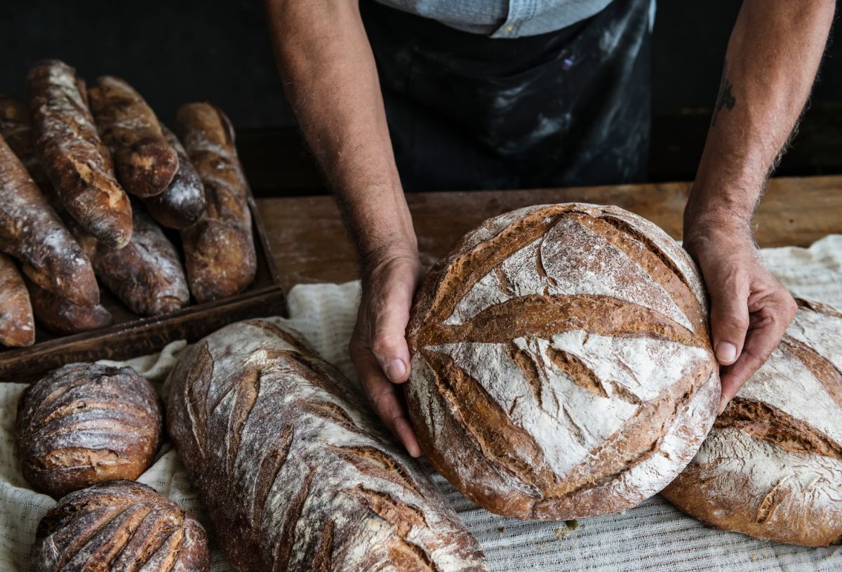 Man holding loaf of bread over table full of loaves of bread