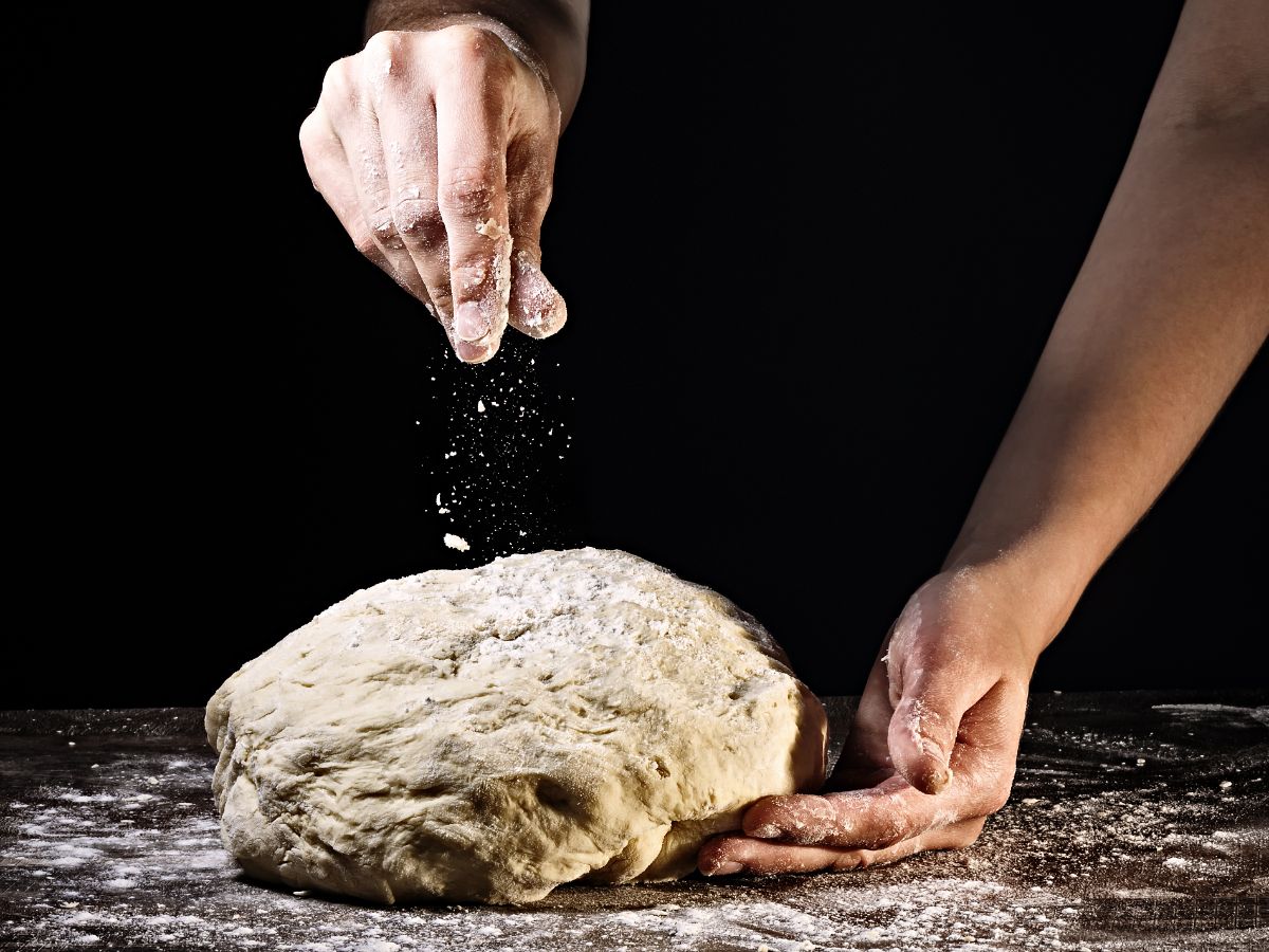 Dough on table being sprinkled by flour in hand