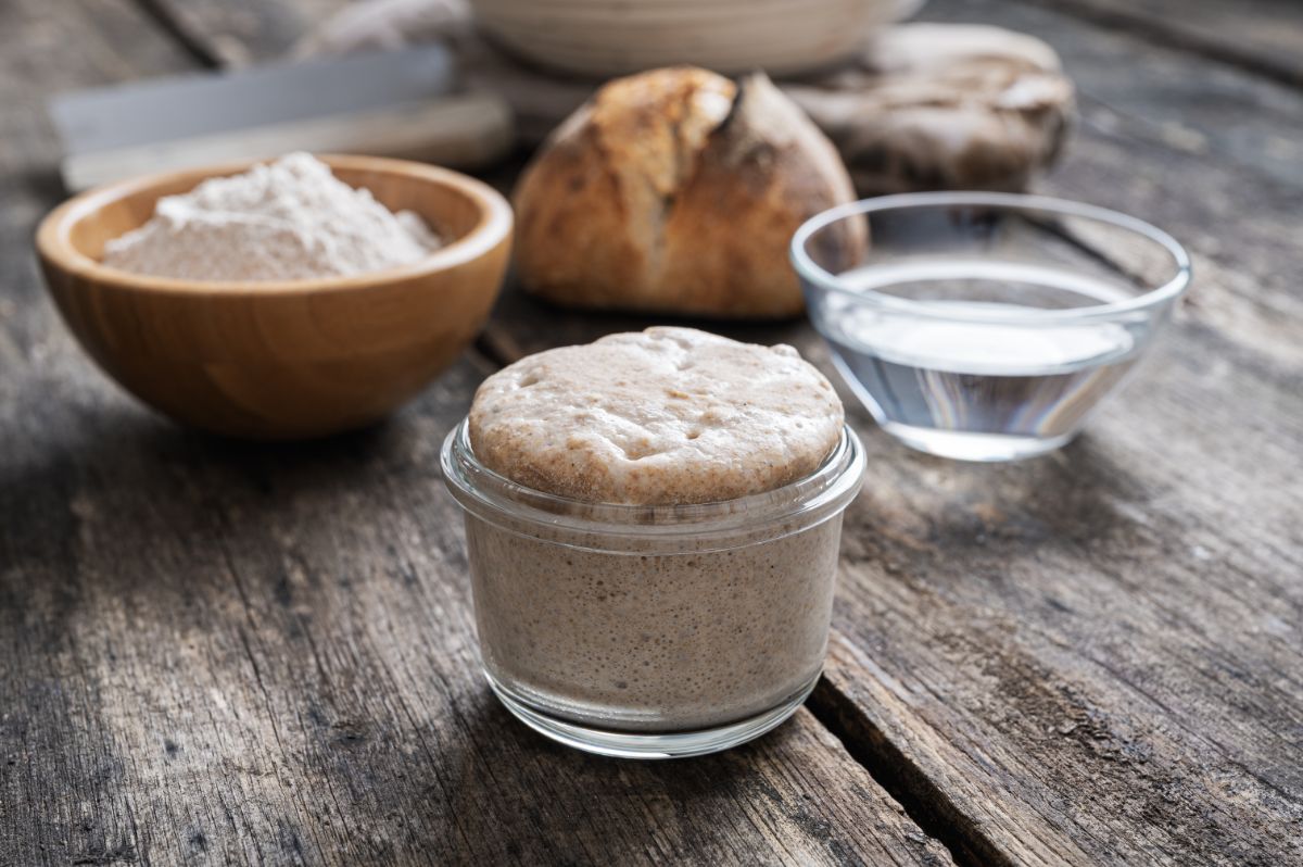 Sourdough starter in glass jar. Bowl of water, bowl of flour and loaf of bread in the background