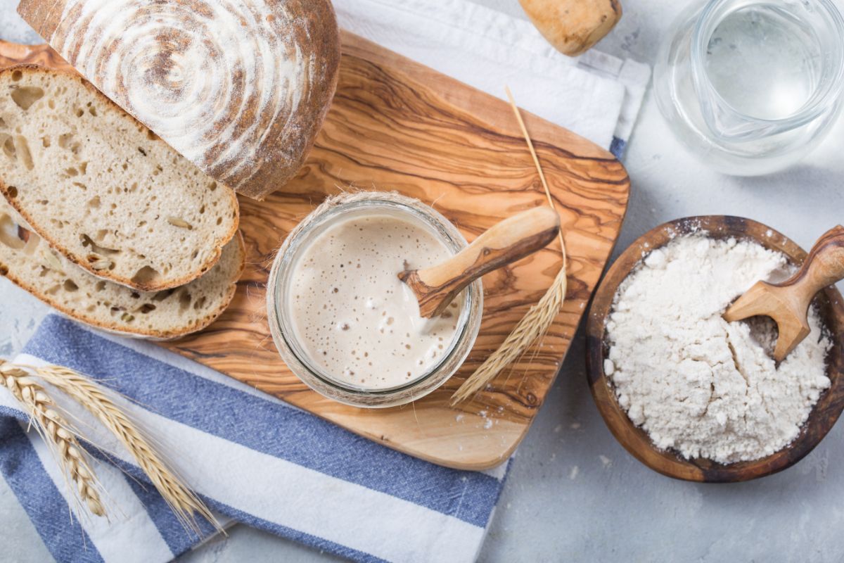Sourdough starter with spoon in glass jar on wooden pad with partialy sliced loaf of bread on table with bowl of  flour and pitcher of water