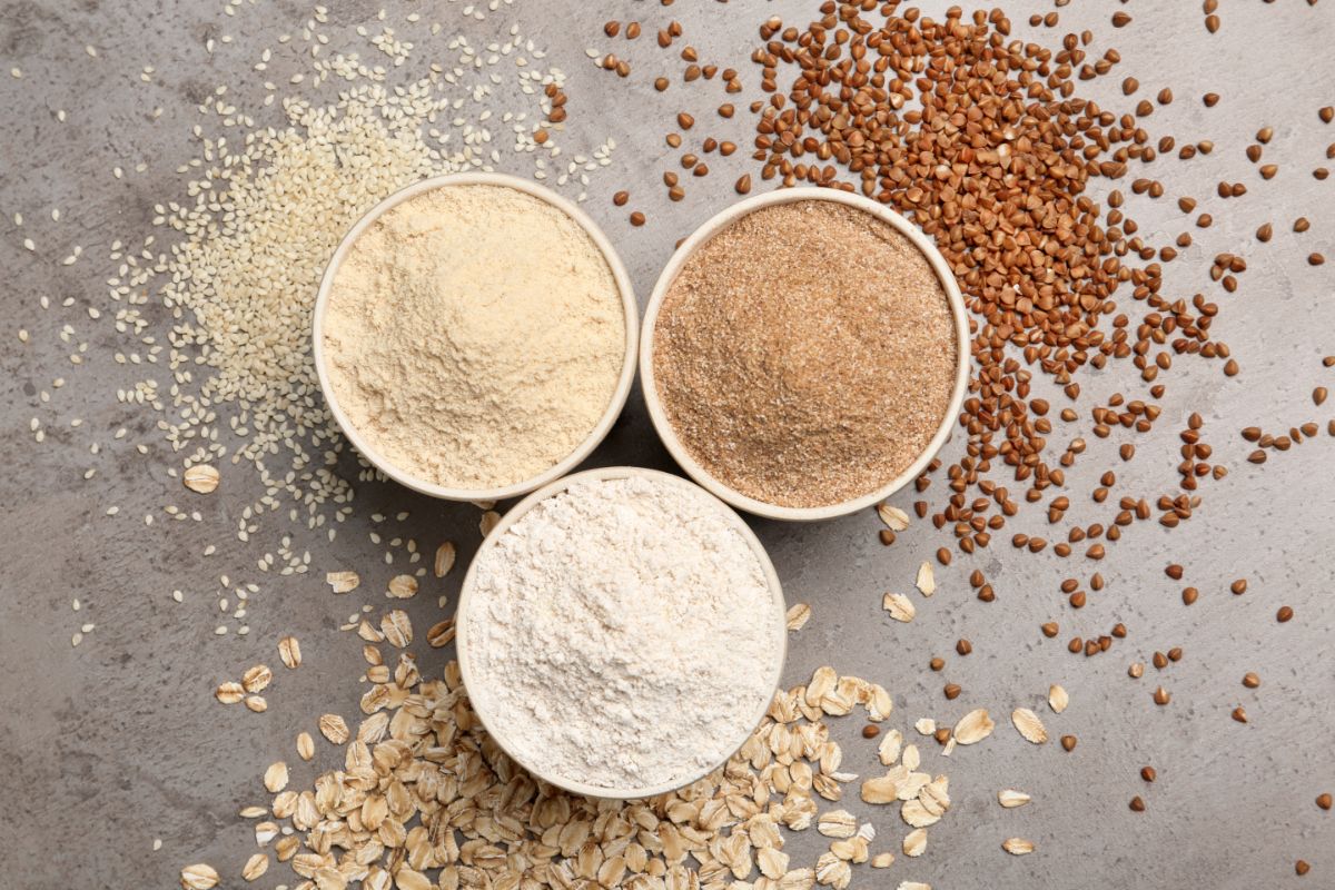 Wheat, nut and oat flour in white bowls with scattered ingredients around on gray table